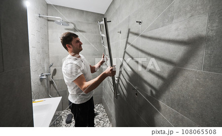 Side view of male plumber hanging electric towel rack dryer on the wall in bathroom. Man installing stainless steel bath towel heater in apartment under renovation. Home renovation concept. 108065508