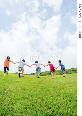 公園の丘を手をつないで駆け上がる小学生たちの後ろ姿 108073794