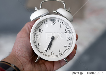 Round white alarm clock in a man's hand close-up. The hands on the clock show half past seven in the morning, time to get up. Retro alarm clock on the table, vintage tone. space for text Round white alarm clock in a man's hand close-up. The hands on the clock show half past seven in the morning, time to get up. Retro alarm clock on the table, vintage tone. space for text 108073893