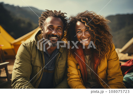 Portrait of happy multiethnic couple in the mountains against the backdrop of a tent camp. Young international family is engaged in a mountain hiking. Active lifestyle, tourism and vacation concept. 108075723