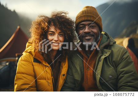 Portrait of happy multiethnic couple in the mountains against the backdrop of a tent camp. Young international family is engaged in a mountain hiking. Active lifestyle, tourism and vacation concept. 108075772