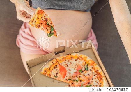A pregnant woman enjoys a slice of pizza, savoring a moment of indulgence while satisfying her craving for a delightful, comforting treat. Excited Pregnant Young Lady Enjoying Pizza Holding Biting 108076637