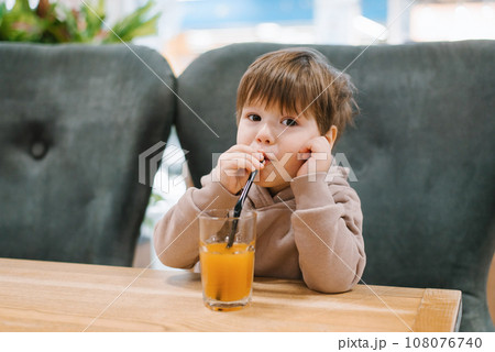 Little boy child drinks juice from a straw while sitting at a table in a cafe 108076740