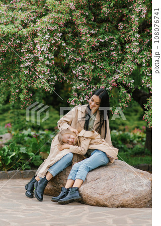 Mother and daughter relax in a spring blooming park, sitting on a stone. The concept of a happy family. Mother and daughter relax in a spring blooming park, sitting on a stone. The concept of a happy family. 108076741