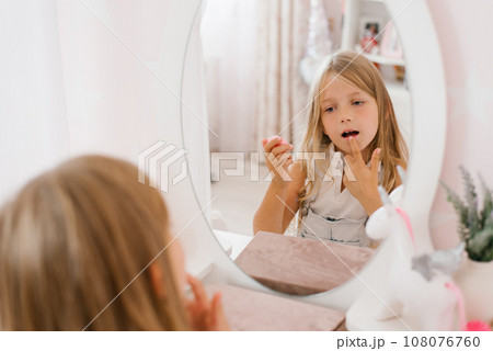 Pretty child girl paints her lips with glitter or balm in front of a mirror in her room 108076760