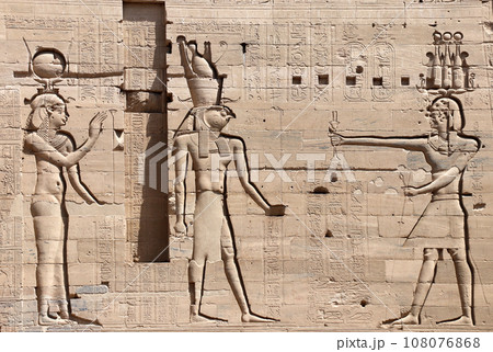 Ancient Egyptian bas-relief on stone wall with figures of the goddess Isis, the god Horus and Pharaoh. Temple of Isis on Agilkia Island (Philae), reservoir of Aswan Low Dam, Egypt Ancient Egyptian bas-relief on stone wall with figures of the goddess Isis, the god Horus and Pharaoh. Temple of Isis on Agilkia Island (Philae), reservoir of Aswan Low Dam, Egypt 108076868