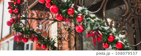 Banner stylish christmas fir branches with red baubles and sparkling garland on front of door at holiday market or restaurant in city street. Winter christmas street decor 108079537
