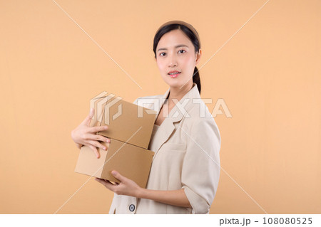 portrait of happy successful confident young asian business woman wearing white jacket holding parcel box isolated on beige studio background, Delivery courier and shipping service concept. portrait of happy successful confident young asian business woman wearing white jacket holding parcel box isolated on beige studio background, Delivery courier and shipping service concept. 108080525