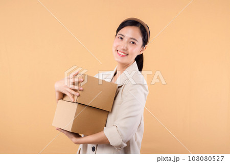 portrait of happy successful confident young asian business woman wearing white jacket holding parcel box isolated on beige studio background, Delivery courier and shipping service concept. 108080527