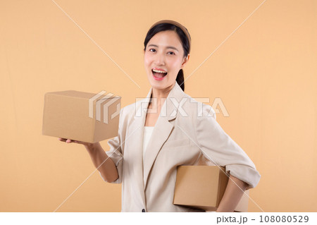 portrait of happy successful confident young asian business woman wearing white jacket holding parcel box isolated on beige studio background, Delivery courier and shipping service concept. 108080529