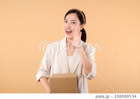 portrait of happy successful confident young asian business woman wearing white jacket holding parcel box isolated on beige studio background, Delivery courier and shipping service concept. 108080534