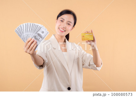 portrait of happy successful confident young asian business woman wearing white jacket holding cash money dollars and credit card standing over beige background. millionaire business, shopping concept 108080573