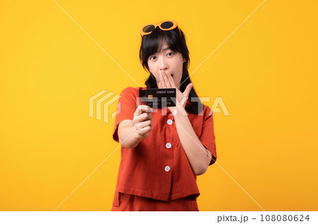 Portrait young beautiful asian woman happy smile dressed in orange clothes showing credit card isolated on yellow background. Pay and purchase shopping payment concept. Portrait young beautiful asian woman happy smile dressed in orange clothes showing credit card isolated on yellow background. Pay and purchase shopping payment concept. 108080624