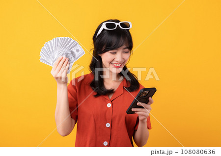 Portrait young beautiful asian woman happy smile dressed in orange clothes showing cash money and smartphone isolated on yellow background. Pay and purchase shopping online payment concept. Portrait young beautiful asian woman happy smile dressed in orange clothes showing cash money and smartphone isolated on yellow background. Pay and purchase shopping online payment concept. 108080636