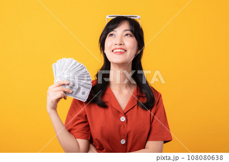 Portrait young beautiful asian woman happy smile dressed in orange clothes dreaming to be rich, success in her life, holding cash money isolated on yellow background. 108080638