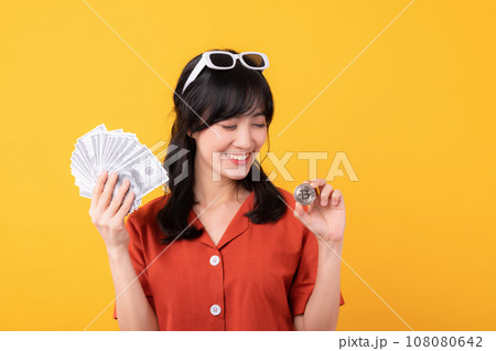 Portrait young beautiful asian woman happy smile dressed in orange clothes holding cash money and crypto bitcoin digital currency celebrate her success isolated on yellow background. Be rich concept. Portrait young beautiful asian woman happy smile dressed in orange clothes holding cash money and crypto bitcoin digital currency celebrate her success isolated on yellow background. Be rich concept. 108080642