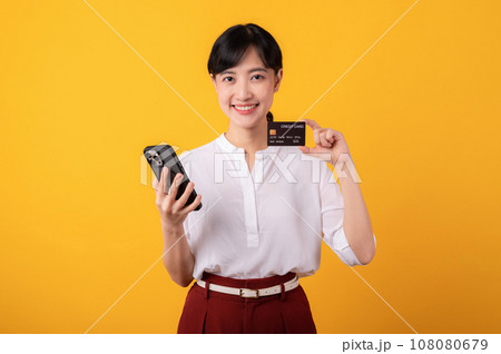 Portrait beautiful young asian woman enterpriser happy smile wearing white shirt and red plants holding credit card and smartphone isolation on yellow background. Online shopping concept. 108080679