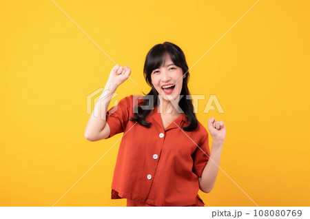 Portrait young beautiful asian woman happy smile dressed in orange clothes showing fist up hand gesture isolated on yellow studio background. Successful hooray celebrate young person concept. 108080769
