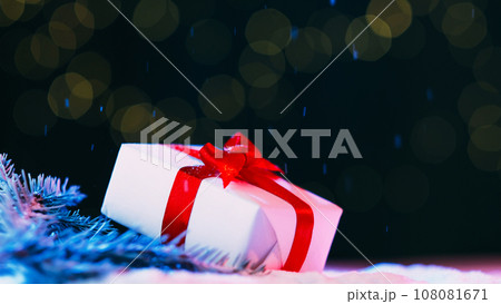 Christmas holiday. Festive gift decor. New Year celebration. Present box wrapped white paper taped red ribbon on winter pine branch snow falling shimmering bokeh lights background. Christmas holiday. Festive gift decor. New Year celebration. Present box wrapped white paper taped red ribbon on winter pine branch snow falling shimmering bokeh lights background. 108081671