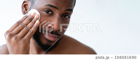 Man care. Face hygiene. Confident satisfied shirtless guy cleansing cheeks facial skin with tonic on cotton pad isolated on white background empty space. Man care. Face hygiene. Confident satisfied shirtless guy cleansing cheeks facial skin with tonic on cotton pad isolated on white background empty space. 108081690