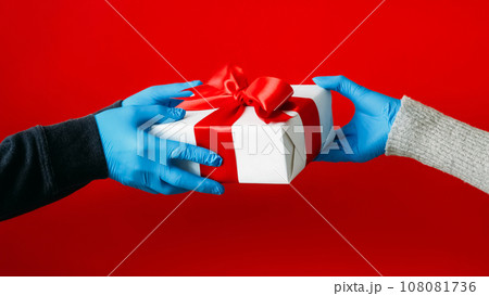 Pandemic gift. Present handing. Unrecognizable human hands in protective gloves holding wrapped box isolated on red background. Pandemic gift. Present handing. Unrecognizable human hands in protective gloves holding wrapped box isolated on red background. 108081736