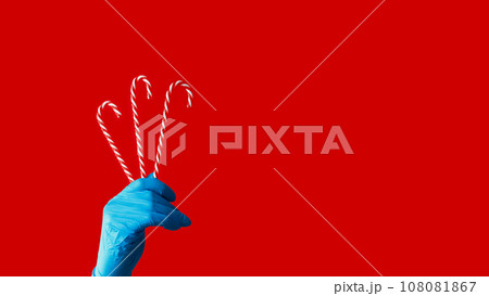 Candy canes. Pandemic holiday. Human hand in blue protective glove holding peppermint sweetmeats isolated on red copy space background. Candy canes. Pandemic holiday. Human hand in blue protective glove holding peppermint sweetmeats isolated on red copy space background. 108081867