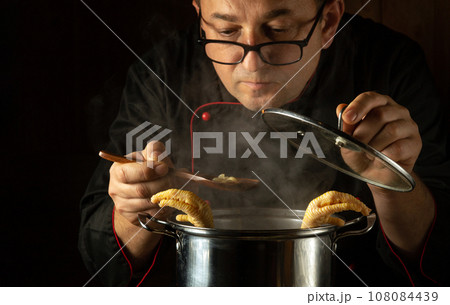 The cook prepares a delicious soup with pasta and chicken in a saucepan. Spoon in the chef's hand. Checking food readiness 108084439