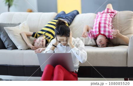 Woman sits on floor with a laptop and covers her ears with her hands. behind her on couch are children playing around. Woman sits on floor with a laptop and covers her ears with her hands. behind her on couch are children playing around. 108085520