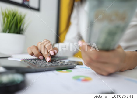 Woman's hand holds dollars and pen with the other hand and types numbers on calculator. Woman's hand holds dollars and pen with the other hand and types numbers on calculator. 108085541
