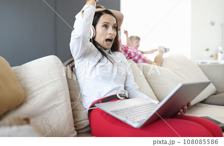 Woman in headphones and with laptops in her hands sits on sofa from behind, children play. 108085586