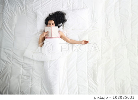 Woman lies on white bed with closed eyes book lies on her face. 108085633