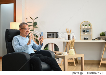 Happy elderly man waving hand while talking online on smartphone with family Happy elderly man waving hand while talking online on smartphone with family 108087472