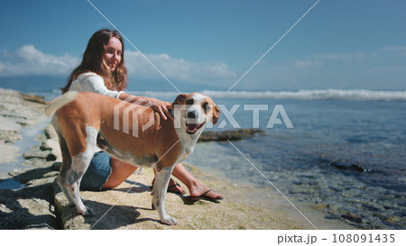 Woman relax with her dogs sitting on ocean coast beach. Young brunette petting the back of red funny pet. Spending time together with best friends. Woman relax with her dogs sitting on ocean coast beach. Young brunette petting the back of red funny pet. Spending time together with best friends. 108091435