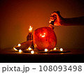 A hand thrusts a knife into a comically carved Halloween pumpkin on a table, surrounded by lanterns 108093498