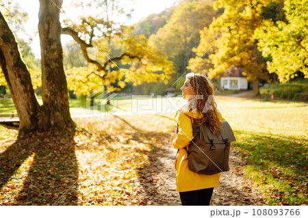 A smiling tourist in a bright coat walks through an autumn park in sunny weather. Travel concept. 108093766