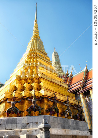 A golden temple spire in Thailand, supported by statues of mythical creatures decorated with colorful mosaic tiles, clear blue sky. A golden temple spire in Thailand, supported by statues of mythical creatures decorated with colorful mosaic tiles, clear blue sky. 108094267