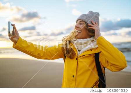 Tourist with phone. Selfie time. Tourist in yellow jacket posing by sea at sunset. Travelling. 108094539