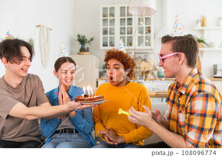 Make a wish. Woman wearing party cap blowing out burning candles on birthday cake. Happy Birthday party. Group of friends wishes girl happy birthday. People celebrating birthday with party at home Make a wish. Woman wearing party cap blowing out burning candles on birthday cake. Happy Birthday party. Group of friends wishes girl happy birthday. People celebrating birthday with party at home 108096774