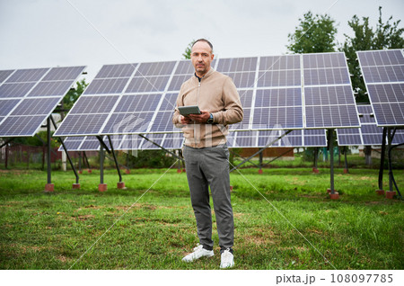 Young investor calculating his future profit from investments in green energy. Alternative energy resources and life improvement. Male with calculator on background solar panel. Young investor calculating his future profit from investments in green energy. Alternative energy resources and life improvement. Male with calculator on background solar panel. 108097785