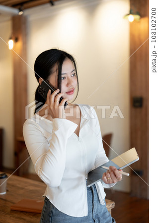 A beautiful Asian woman is calling or talking with someone on her phone while standing in a cafe. 108102700