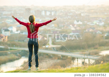 Young woman standing outdoors raising her hands enjoying city view. Relaxing, freedom and wellness concept. 108104562