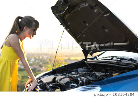 Young woman driver standing near her car with popped hood having engine problems. 108104584