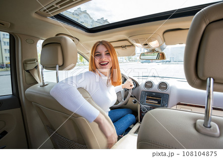 Wide angle view of young redhead woman driver driving a car backwards looking behind. 108104875
