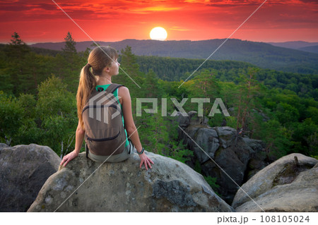 Tired hiker girl relaxing on rocky mountain top enjoying evening nature during travelling on wilderness trail. Lonely woman traveler traversing high hilltop route. Healthy lifestyle concept 108105024