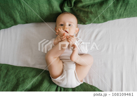Cute baby is lying on a green and white bedspread on the bed. A smiling child having his feet in a sunny bedroom. Cute baby is lying on a green and white bedspread on the bed. A smiling child having his feet in a sunny bedroom. 108109461