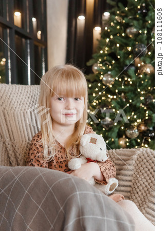 A little blonde girl is sitting in an armchair in an interior decorated in a New Year's style. A child holds a teddy bear in his hands. 108111608
