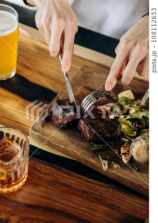 Close up of hands holding knife and fork is cutting grilled ribeye steak on wooden cutting board Close up of hands holding knife and fork is cutting grilled ribeye steak on wooden cutting board 108112653