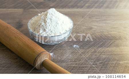 Wooden rolling pin and bowl with flour on wooden table, front view. Baking concept. Wooden rolling pin and bowl with flour on wooden table, front view. Baking concept. 108114654