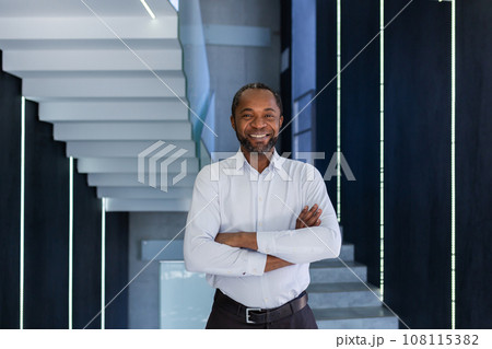 Mature african american businessman with crossed arms smiling and looking at camera inside office building, satisfied and successful man at work in shirt. Mature african american businessman with crossed arms smiling and looking at camera inside office building, satisfied and successful man at work in shirt. 108115382