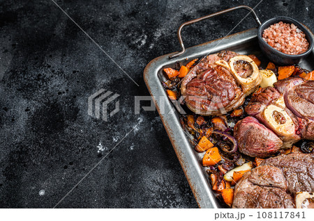 Italian braised veal shank steak Ossocbuco with vegetable sauce on steel tray. Black background. Top view. Copy space Italian braised veal shank steak Ossocbuco with vegetable sauce on steel tray. Black background. Top view. Copy space 108117841